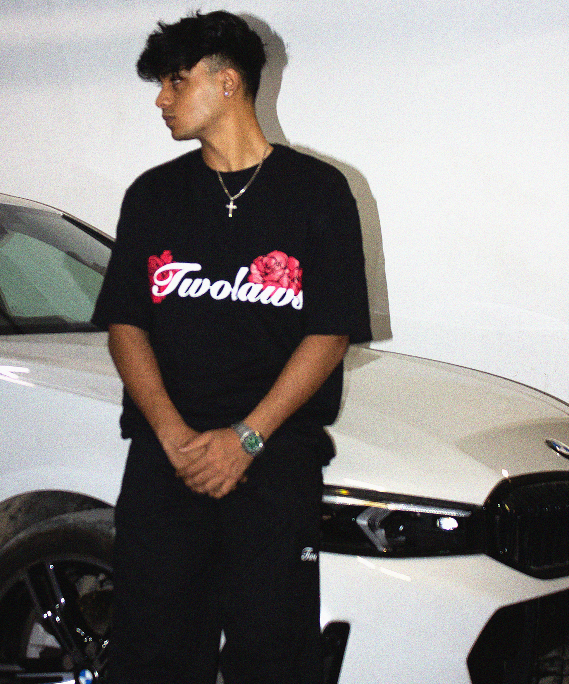 Model leaning on a supercar wearing the TWOLAWS Rose T-Shirt with script font and rose design.